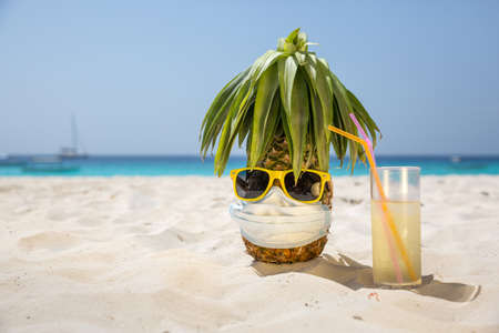 A ripe and juicy pineapple in a protective mask and yellow sunglasses stands on the beach, on white sand, next to a glass with a cool drink, in the background the azure sea and sailboatsの写真素材