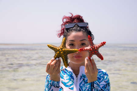 a beautiful girl in a swimsuit stands against the background of the sea and holds starfish in front of her faceの写真素材