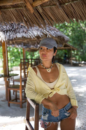 A beautiful girl in jeans shorts, a baseball cap and a yellow shirt stands under a cane canopy...