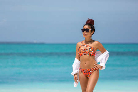 Beautiful sexy brunette girl in a bikini with a leopard print and a white shirt and sunglasses stands on the beach against the background of the azure oceanの写真素材