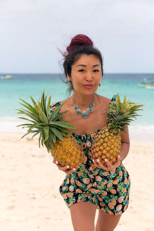 Beautiful happy asian woman wearing pineapple print clothes holding pineapples in front of her on the seashoreの写真素材