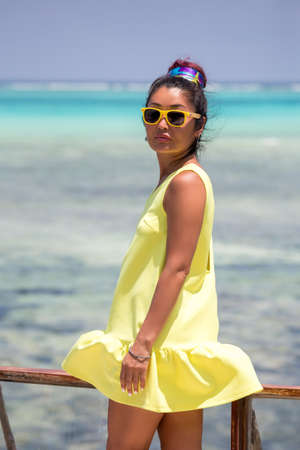 A beautiful tanned girl in a short yellow dress and yellow glasses stands on the terrace against the background of the azure oceanの写真素材