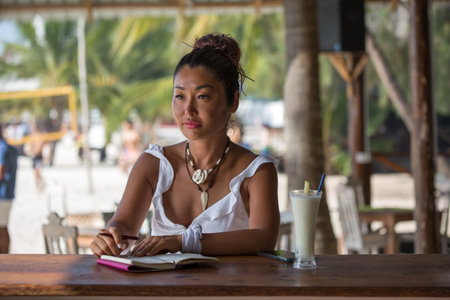 A beautiful tanned girl in a white dress sits at the bar on the terrace with a notebook and a pen and drinks a cocktailの写真素材