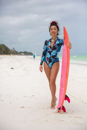 A beautiful brunette girl, a kitesurfer with a board in a bikini stands on the beach against the...