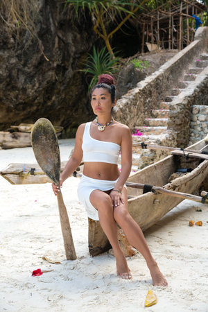 A beautiful girl model in a white beach suit with an oar in her hands sits on an old fishing boat near the stone stepsの写真素材