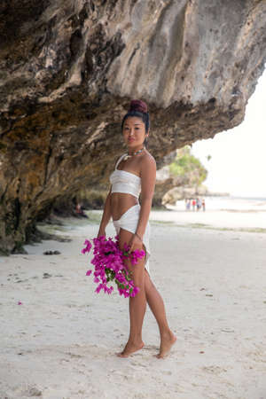 A beautiful girl model in a white beach suit with a bouquet of lilac flowers stands by the cliff against the background of the seaの写真素材