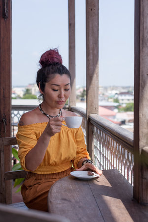 A beautiful brunette girl of Asian appearance in an orange blouse and terracotta trousers drinks coffee on the balconyの写真素材
