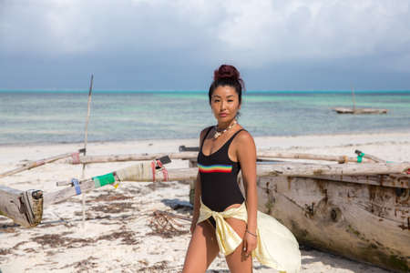A beautiful girl in a black swimsuit with an LGBT logo and a yellow shirt stands near a fishing catamaran on the beachの写真素材