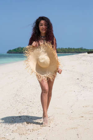 A beautiful brunette girl stands naked on the beach, hiding behind a huge straw hat. In the background, a deserted white beachの写真素材