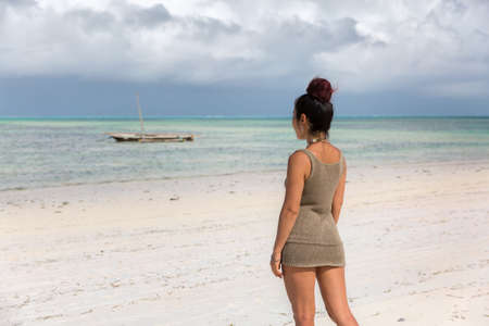 a slender beautiful girl in a short dress stands by the ocean and looks into the distance, the sky is covered with cloudsの写真素材