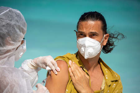A doctor in a protective suit gives a covid-19 vaccine to a young attractive European man in a...