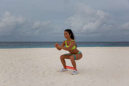 A young attractive sexy girl in a sports uniform, in a bikini, does physical exercises on the beach by the sea, at sunset. Background blue sea and cloudy skyの写真素材