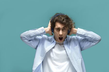 Portrait of a young beautiful man wearing white t-shirt and blue shirt in headphones covers ears with hands shouting from noise and looking on camera over blue backgroundの写真素材
