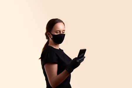 woman in black dress face mask and latex gloves holding mobile phone over beige backgroundの写真素材