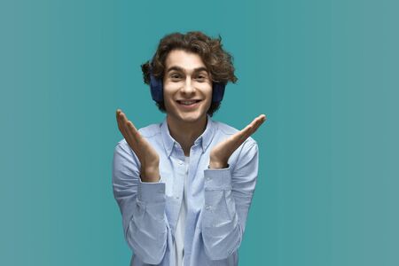 Very cute. Portrait of a young beautiful man wearing white t-shirt and blue shirt in headphones smile holds palms upward to his face and looking at camera over blue backgroundの写真素材