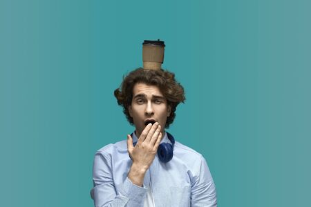 Morning distraction. Portrait of a young beautiful yawninig man wearing white t-shirt and blue shirt in headphones with hot drink cup in haad over blue backgroundの写真素材