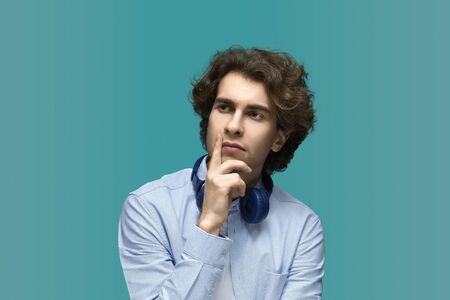 Let me think. Portrait of a young beautiful man wearing white t-shirt and blue shirt in blue headphones.Thought and holds a fist at the chin over blue backgroundの写真素材