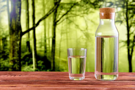 A glass and a jug full of drinking water on a wooden table over natural green forest background