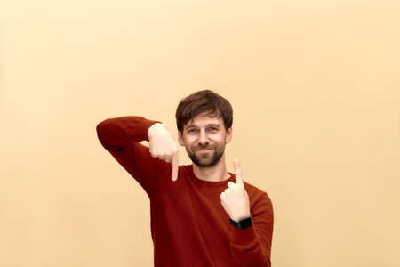 Photo of young man with beard wearing sweater, points at different sides on yellow wall as shows something pleasant, has smiling look, advertises product.の写真素材