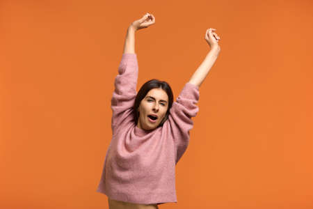 I woke up. Beautiful woman in pink sweater stretches and yawns as being tired and needs good rest after hard work, gets up early in morning, isolated on orange background.の写真素材