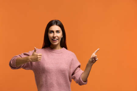 Its a great idea. Indoor shot of cheerful beautiful female shows blank space right, keeps thumb up advertises something. wears in pink knitted sweater isolated over orange backgroundの写真素材