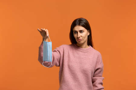 Tired of coronavirus. Dissatisfied young female holds and looking at medical mask. looks sadly. give up of using it. wears in pink knitted sweater isolated over orange backgroundの写真素材