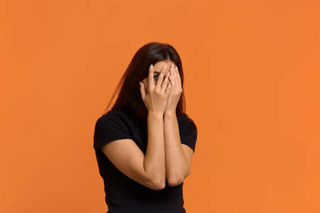 Scared caucasian woman in black t-shirt peeps through fingers, hides face with hands, afraids of...