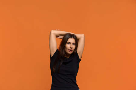 Relaxed carefree caucasian woman in black t-shirt keeps hands behind head, has positive expression,...