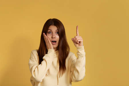 wow price. Amazed surprised caucasian woman in yellow hoodie with shocking expression indicates with fore finger upwards, being stunned of something.Isolated over a yellow backgroundの写真素材