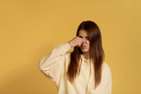 Young caucasian woman in a yellow hoodie covers nose with hand, smells something awful, pinches nose, frowns in displeasure, sees pile of garbage, feels bad odour, isolated over yellow backgroundの写真素材