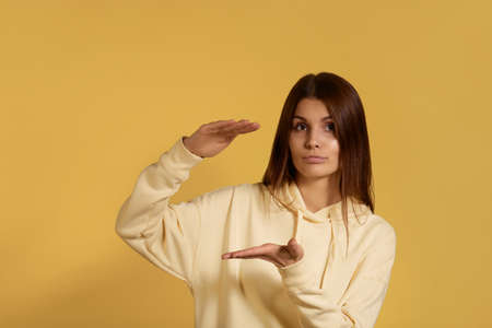 Young caucasian woman in a yellow hoodie shows big size of received present, shapes height of gift...