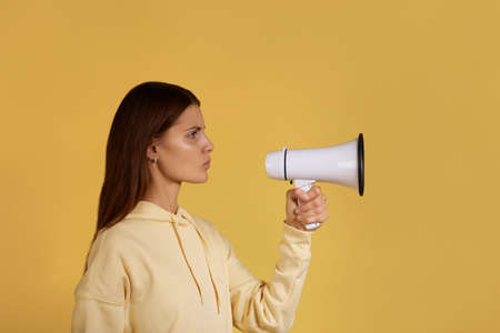 Protest. Young calm caucasian woman in yellow hoodie stands quiet holding a megaphone, ready to speak out at loudspeaker, disagrees, shows her opinion, isolated on yellow backgroundの写真素材