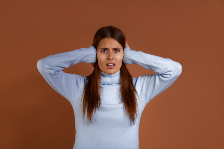 Switch it off. Irritated young european woman wearing blue sweater is annoyed by loud noise, closes ears with hands, frowns, looks straight with disgust, has mouth open isolated on brown backgroundの写真素材