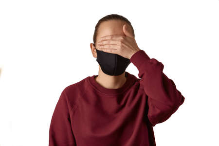 Tired young european woman wears black face mask and burgundy sweatshirt, closes eyes with her hand...