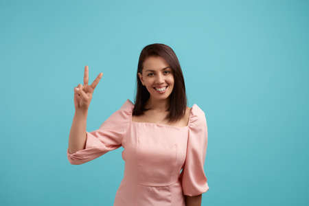 Joyful beautiful woman in pink dress shows peace sign and welcomes everyone smiles positively, makes...