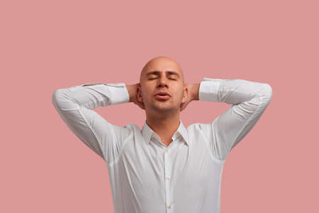 I can relax everywhere. Horizontal photo shot of relaxed carefree bald guy in white shirt, keeps hands behind head, has calm expression, isolated over pink backgroundの写真素材