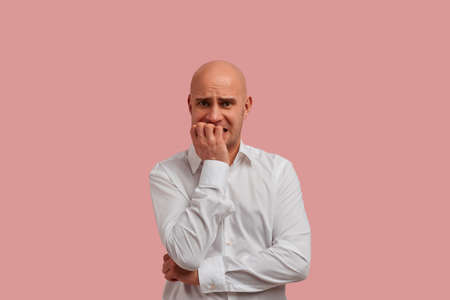 Can i tomorrow. Horizontal portrait of nervous bald man with bristle, bites finger nails anxiously, worried of go to the boss for an audience. Dressed in white shirt. Isolated on pink backgroundの写真素材