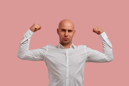 Strong bald man with bristle posing raises hands and shows muscles, his strength and power, feels proud about his achievements in gym. Dressed in white shirt, isolated over pink backgroundの写真素材