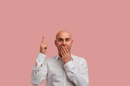 What a sale. Incredible prices. Shocked bald guy with bristle in white shirt covers mouth not speak, points to up with forefinger, shows something amazed. Isolated over pink backgroundの写真素材