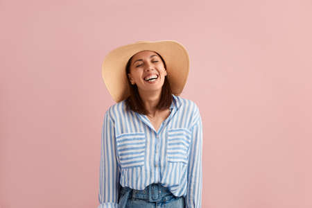 Cheerful relaxed woman with dark hair laughs, closes eyes in excitement, hands down, feels carefree, wears straw hat, striped shirt, jeans with belt, on pink background. Summer emotions conceptの写真素材