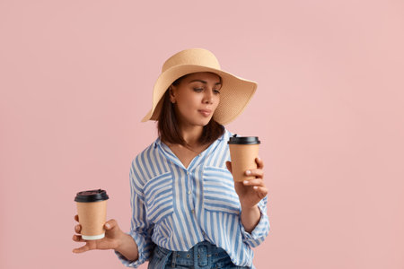 Peaceful brunette is holding two cups of takeaway morning coffee on the road, cannot decide, feels smell, wears straw hat, striped shirt, jeans with belt, on pink background. Summer emotions conceptの写真素材