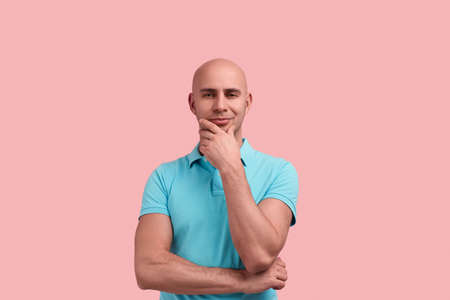 Handsome homosexual bald man with bristle in doubt, does not believe you, makes decision, touches chin, keeps palm under elbow, gay friendly, wears blue polo shirt, poses over pink backgroundの写真素材