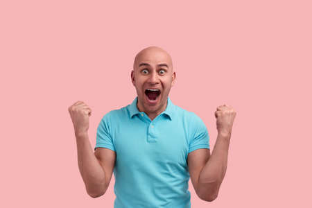 Happy surprised bald homosexual man with bristle won a prize, screams in excitement, has mouth wide open, clenches fists, gay friendly, wears blue polo shirt, poses over pink backgroundの写真素材