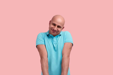 Embarrassed kind bald homosexual man with bristle is touched and impressed with gift, closes eyes in pleasure, shrugs, gay friendly, wears blue polo shirt, poses over pink backgroundの写真素材