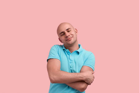 Love myself. Glad bald homosexual man with bristle hugs himself, keeps eyes closed, smiles with pleasure.の写真素材