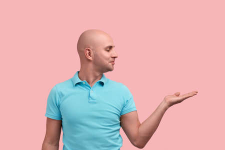 Pleased bald unshaven homosexual man shows a product or offer with his palm, looks aside with proud and pleasure.の写真素材