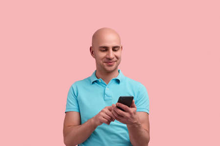 Pleased bald unshaven homosexual man is scrolling feed, shopping online, chatting in date app, enjoys communication.の写真素材