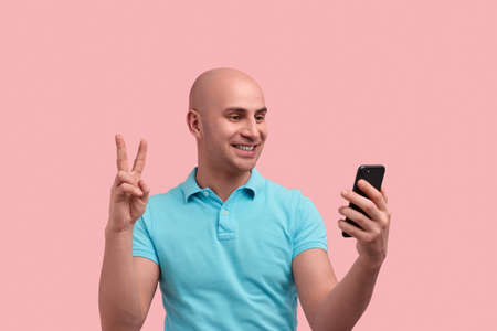 Cheerful smiling bald unshaven homosexual man shows victory or peace sign, looking at mobile phone...