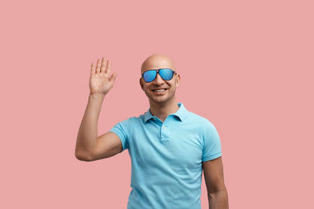 Cheerful smiling bald unshaven homosexual man waves hand, says hello, smiles, raises palm, happy to meet friends.の写真素材