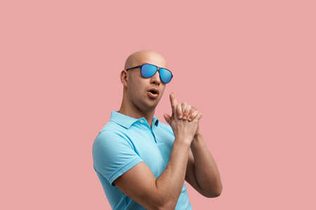 Handsome bald unshaven homosexual man fakes gun with hands, makes shooting gesture, achieves goal, keeps mouth open.の写真素材